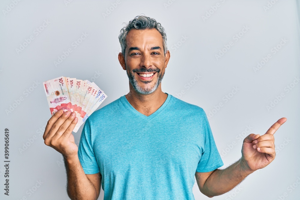 Middle age grey-haired man holding colombian pesos smiling happy pointing with hand and finger to the side Middle age grey-haired man holding colombian pesos smiling happy pointing with hand and finger to the side