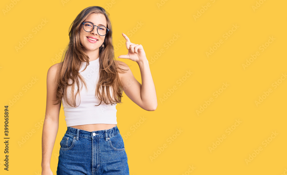 Beautiful caucasian young woman wearing casual clothes and glasses smiling and confident gesturing with hand doing small size sign with fingers looking and the camera. measure concept. Beautiful caucasian young woman wearing casual clothes and glasses smiling and confident gesturing with hand doing small size sign with fingers looking and the camera. measure concept.