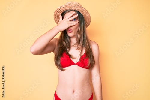 Beautiful young caucasian woman wearing bikini and hat peeking in shock covering face and eyes with hand, looking through fingers afraid