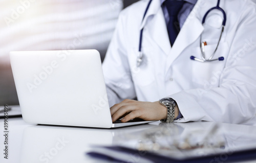 Unknown male doctor sitting and working with laptop in a darkened clinic, glare of light on the background, close-upof hands