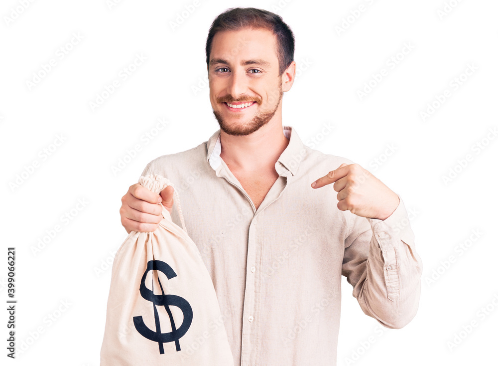 Young handsome caucasian man holding money bag with dollar symbol pointing finger to one self smiling happy and proud Young handsome caucasian man holding money bag with dollar symbol pointing finger to one self smiling happy and proud
