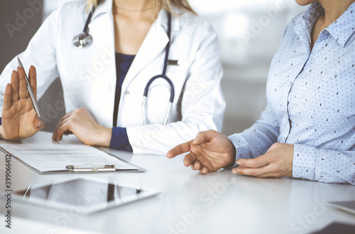 Unknown woman- doctor is listening to her patient, while sitting together at the desk in the cabinet in a clinic. Female physician with a stethoscope is writing at clipboard, close up. Perfect medical