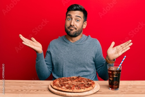 Handsome hispanic man eating tasty pepperoni pizza clueless and confused with open arms, no idea and doubtful face.