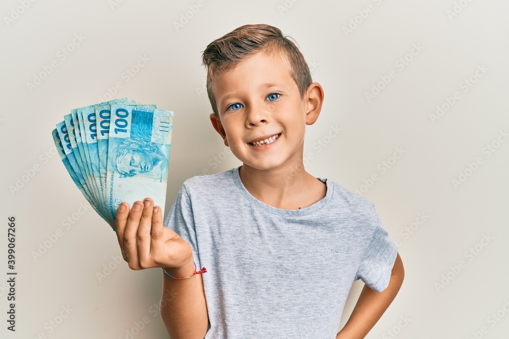 Adorable caucasian kid holding 100 brazilian real banknotes looking positive and happy standing and smiling with a confident smile showing teeth Adorable caucasian kid holding 100 brazilian real banknotes looking positive and happy standing and smiling with a confident smile showing teeth