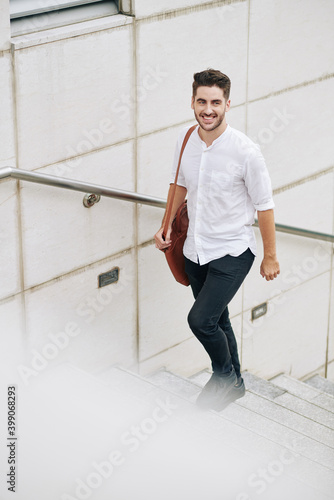 Cheerful handsome young bearded man in cotton white shirt walking up the stairs