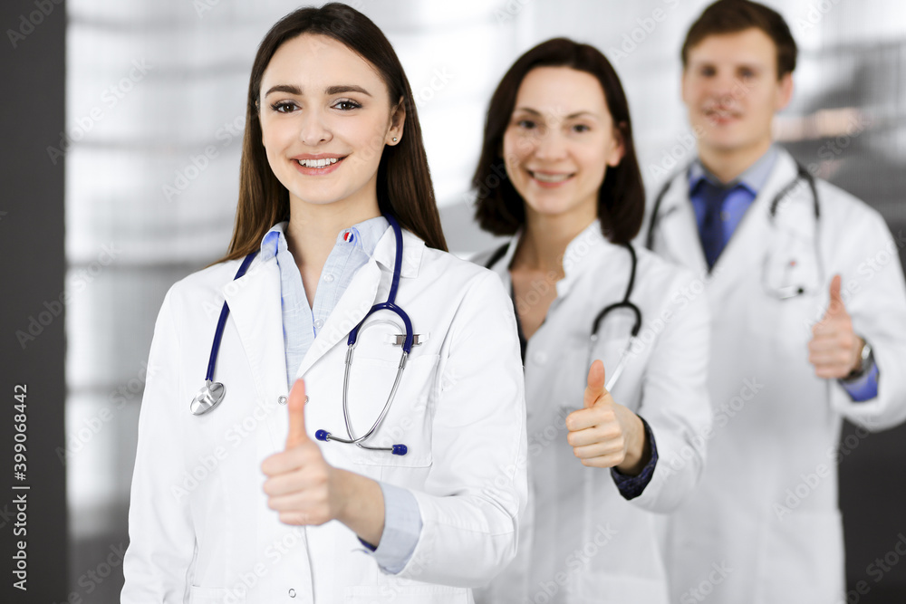 Group of young cheerful doctors is standing as a team with thumbs up in a hospital office and is ready to help patients. Medical help, insurance in health care, best disease treatment and medicine Group of young cheerful doctors is standing as a team with thumbs up in a hospital office and is ready to help patients. Medical help, insurance in health care, best disease treatment and medicine