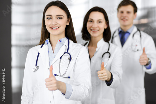 Group of young cheerful doctors is standing as a team with thumbs up in a hospital office and is ready to help patients. Medical help, insurance in health care, best disease treatment and medicine