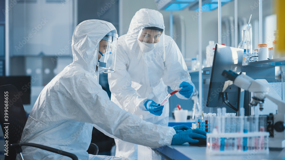 Medical Laboratory: Team of Microbiology Scientists Wearing Sterile Coveralls, Face Shields and Masks Talk, Use Computer to Analyse Test Tube Blood Samples and Develop Vaccine, Drugs and Antibiotics Medical Laboratory: Team of Microbiology Scientists Wearing Sterile Coveralls, Face Shields and Masks Talk, Use Computer to Analyse Test Tube Blood Samples and Develop Vaccine, Drugs and Antibiotics