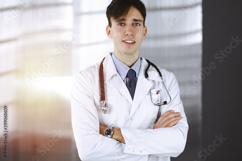 Cheerful smiling man-doctor standing with arms crossed in clinic. Perfect medical service with young smart physician in hospital. Medicine concept