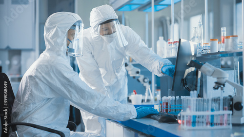 Medical Laboratory: Team of Microbiology Scientists Wearing Sterile Coveralls, Face Shields and Masks Talk, Use Computer to Analyse Test Tube Blood Samples and Develop Vaccine, Drugs and Antibiotics