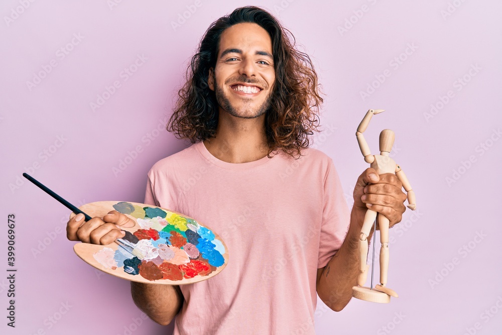 Young handsome man with long hair holding painter palette and art manikin smiling with a happy and cool smile on face. showing teeth. Young handsome man with long hair holding painter palette and art manikin smiling with a happy and cool smile on face. showing teeth.