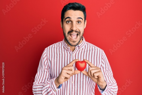 Young hispanic man holding heart celebrating crazy and amazed for success with open eyes screaming excited.