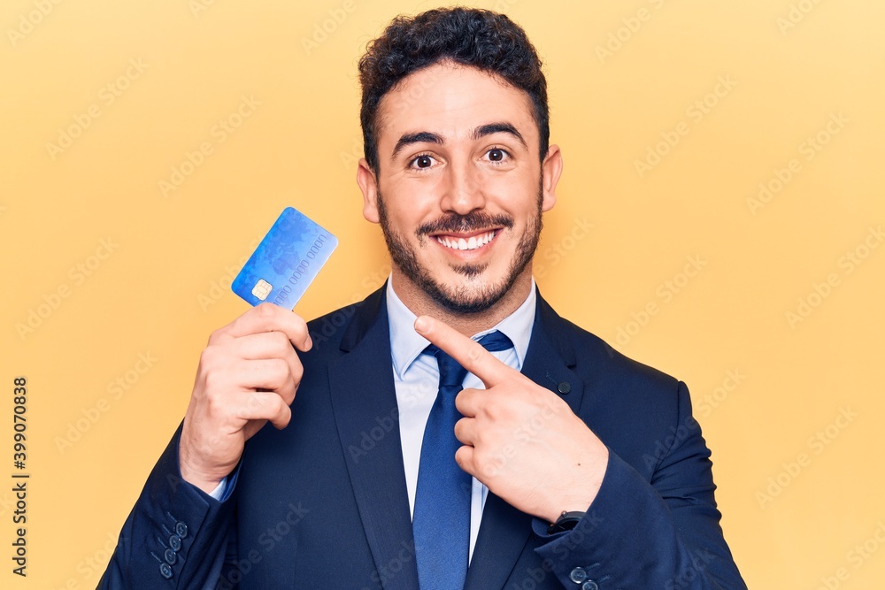 Young hispanic man wearing suit holding credit card smiling happy pointing with hand and finger Young hispanic man wearing suit holding credit card smiling happy pointing with hand and finger