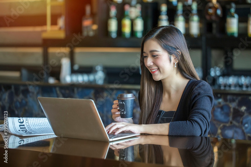 Beautiful Asian businesswoman sitting at work, reading the newspaper and reading information online on her laptop, drinking coffee in the morning at a coffee shop.