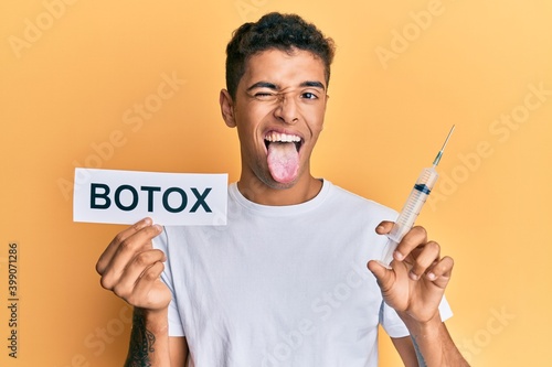 Young handsome african american man holding botox banner and needle sticking tongue out happy with funny expression.