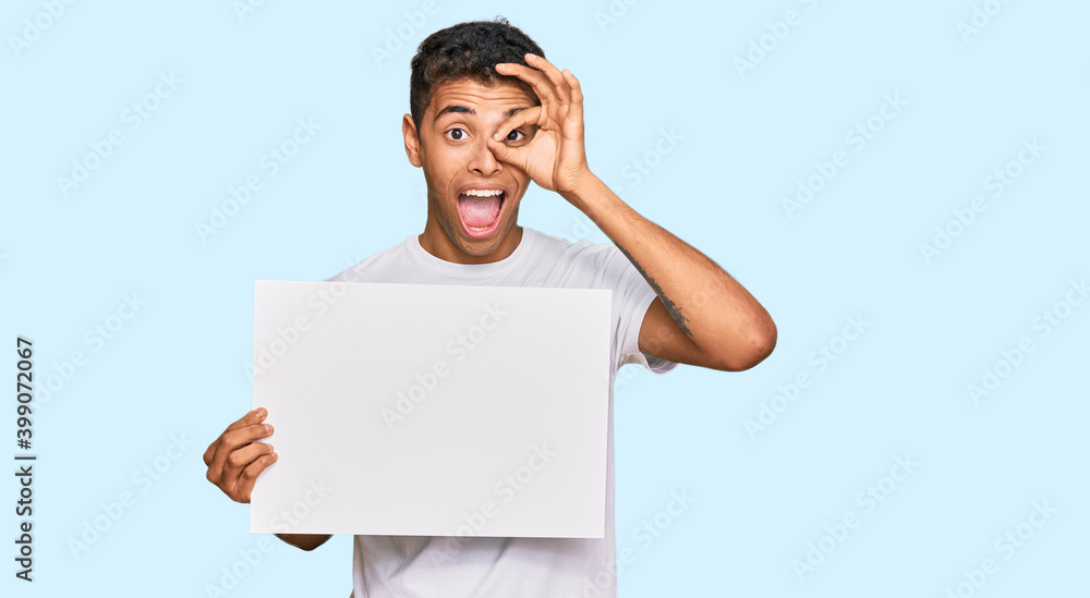 Young handsome african american man holding blank empty banner smiling happy doing ok sign with hand on eye looking through fingers Young handsome african american man holding blank empty banner smiling happy doing ok sign with hand on eye looking through fingers