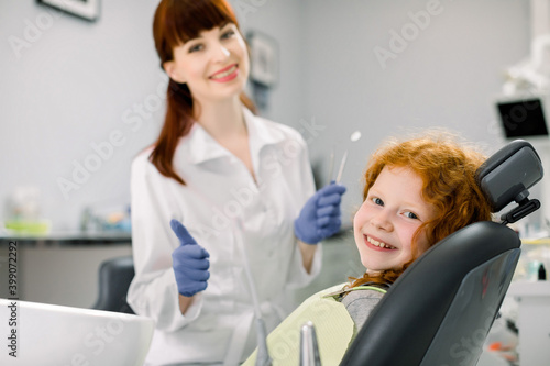 Side view of cheerful Caucasain girl with curly red hair, lying on dentist chair, looking at camera and smiling. Professional female dentist on the background, holds dental tool and shows thumbs up