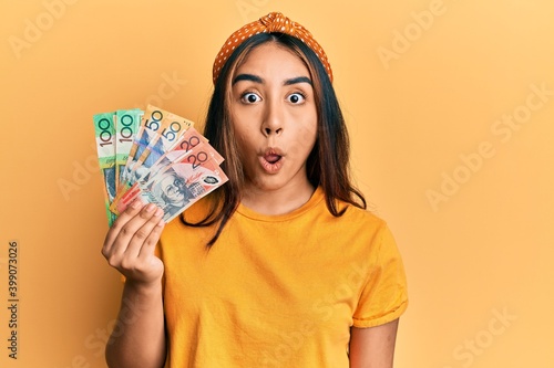 Young latin woman holding australian dollars banknotes scared and amazed with open mouth for surprise, disbelief face