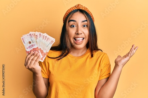 Young latin woman holding colombian pesos celebrating achievement with happy smile and winner expression with raised hand