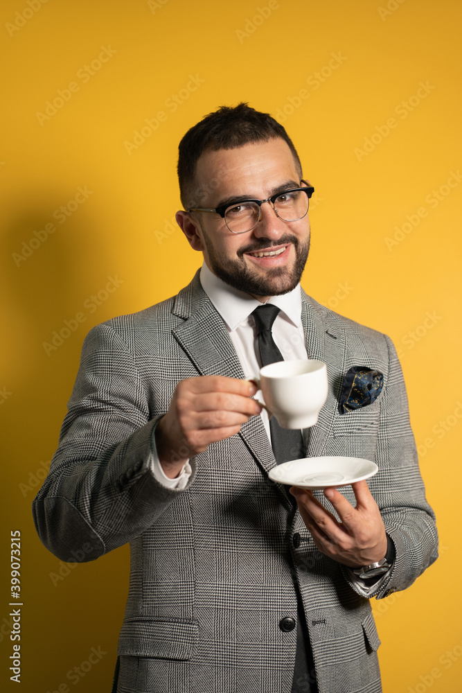 Cheerful young handsome bearded man in eye glasses in formal wear holding a cup of coffee isolated on yellow background. Cheerful young handsome bearded man in eye glasses in formal wear holding a cup of coffee isolated on yellow background.