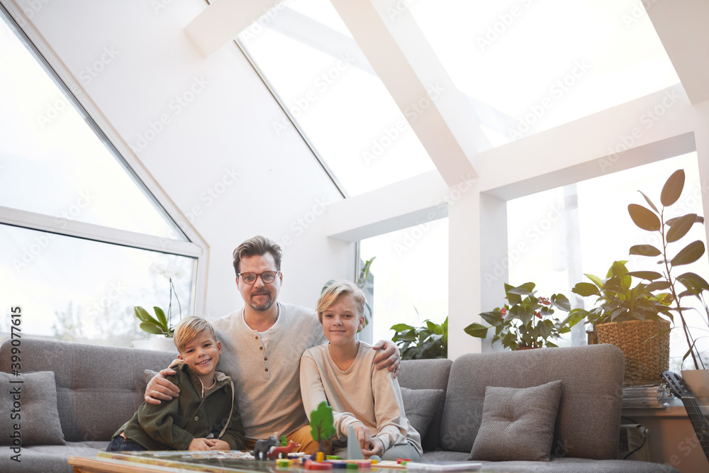 Wide angle portrait of loving father embracing two sons and looking at camera while sitting on sofa together in modern home interior, copy space Wide angle portrait of loving father embracing two sons and looking at camera while sitting on sofa together in modern home interior, copy space
