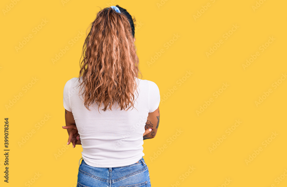 Young hispanic woman with tattoo wearing casual white tshirt standing backwards looking away with crossed arms