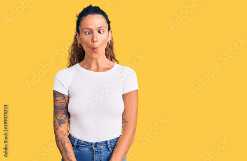 Young hispanic woman with tattoo wearing casual white tshirt making fish face with lips, crazy and comical gesture. funny expression.