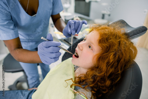 Side close up view of curly red haired girl lying on dentist chair with open mouth while female African doctor in blue uniform makes dental checkup with tools. Concept of teeth treatment and care.