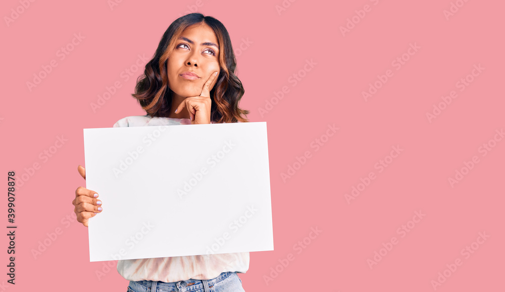 Young beautiful mixed race woman holding cardboard banner with blank space serious face thinking about question with hand on chin, thoughtful about confusing idea Young beautiful mixed race woman holding cardboard banner with blank space serious face thinking about question with hand on chin, thoughtful about confusing idea