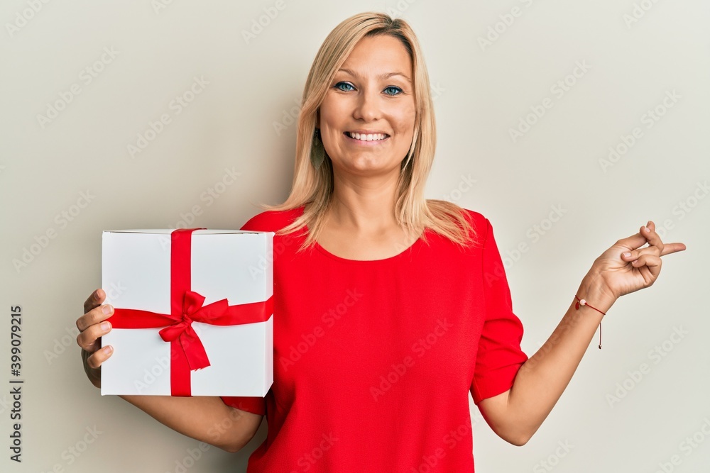 Beautiful middle age blonde woman holding gift smiling happy pointing with hand and finger to the side Beautiful middle age blonde woman holding gift smiling happy pointing with hand and finger to the side