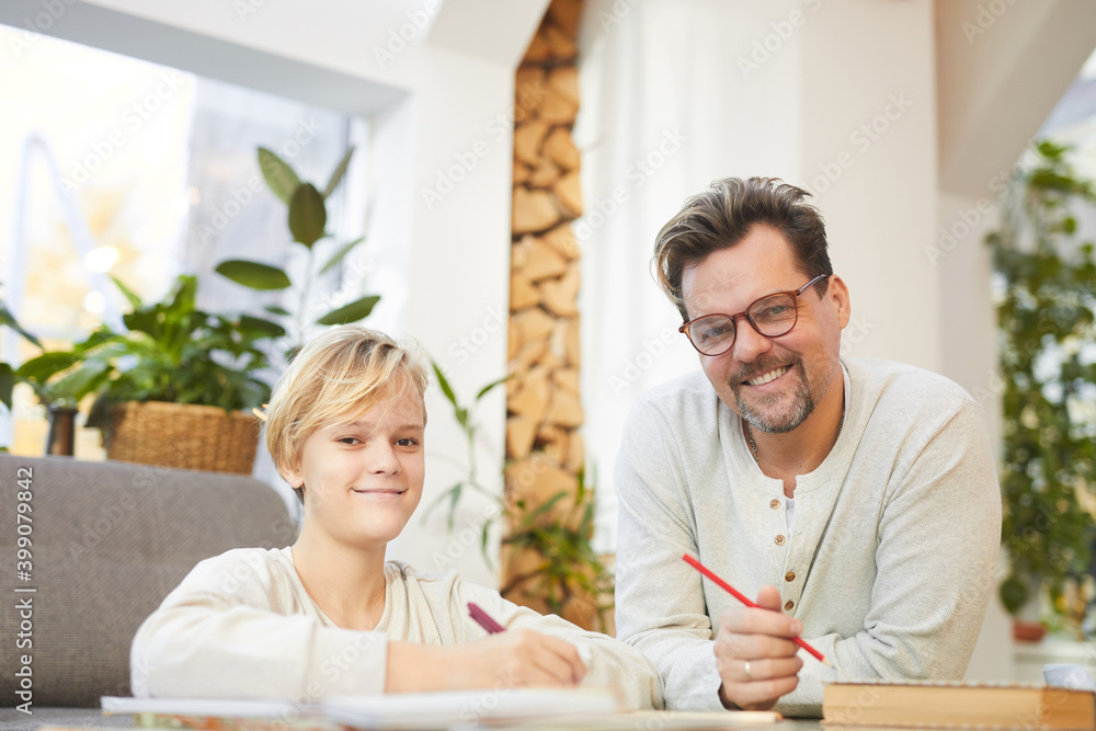 Portrait of modern happy father and son smiling at camera while doing homework together at table in home interior, copy space Portrait of modern happy father and son smiling at camera while doing homework together at table in home interior, copy space