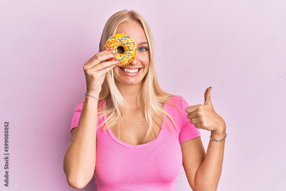Young blonde girl holding donut smiling happy and positive, thumb up doing excellent and approval sign Young blonde girl holding donut smiling happy and positive, thumb up doing excellent and approval sign