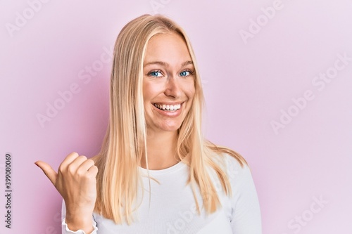 Young blonde girl wearing casual clothes smiling with happy face looking and pointing to the side with thumb up.
