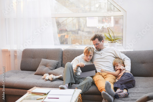Wide angle portrait of modern mature father lying on sofa with two sons and watching videos via tablet together, copy space