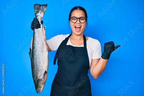 Young beautiful asian girl fishmonger selling fresh raw salmon pointing thumb up to the side smiling happy with open mouth photo