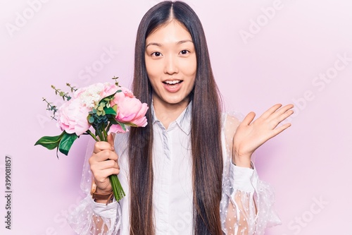 Young beautiful chinese woman holding pink bouquet of flowers celebrating achievement with happy smile and winner expression with raised hand