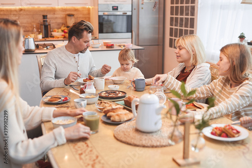 Wide angle portrait of big happy family enjoying breakfast together in kitchen , focus on father helping cute baby girl, copy space