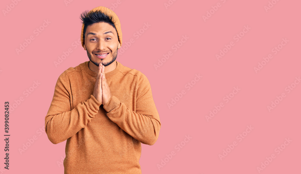 Fototapeta premium Handsome latin american young man wearing winter sweater and wool hat praying with hands together asking for forgiveness smiling confident.