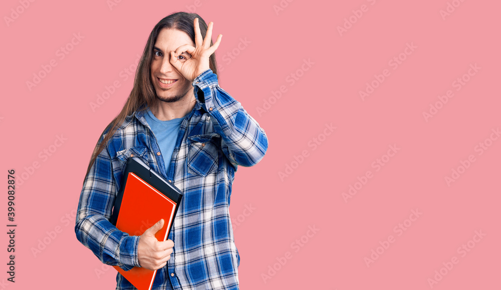 Young adult man with long hair holding collage book smiling happy doing ok sign with hand on eye looking through fingers Young adult man with long hair holding collage book smiling happy doing ok sign with hand on eye looking through fingers