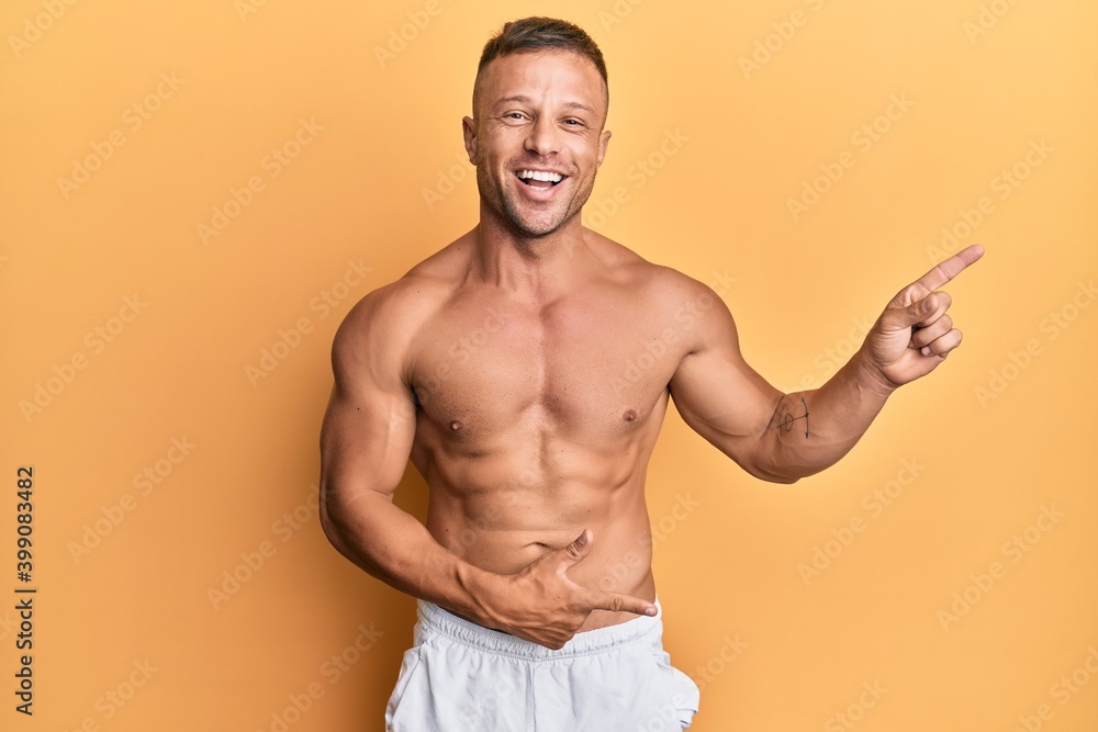 Handsome muscle man standing shirtless smiling and looking at the camera pointing with two hands and fingers to the side.