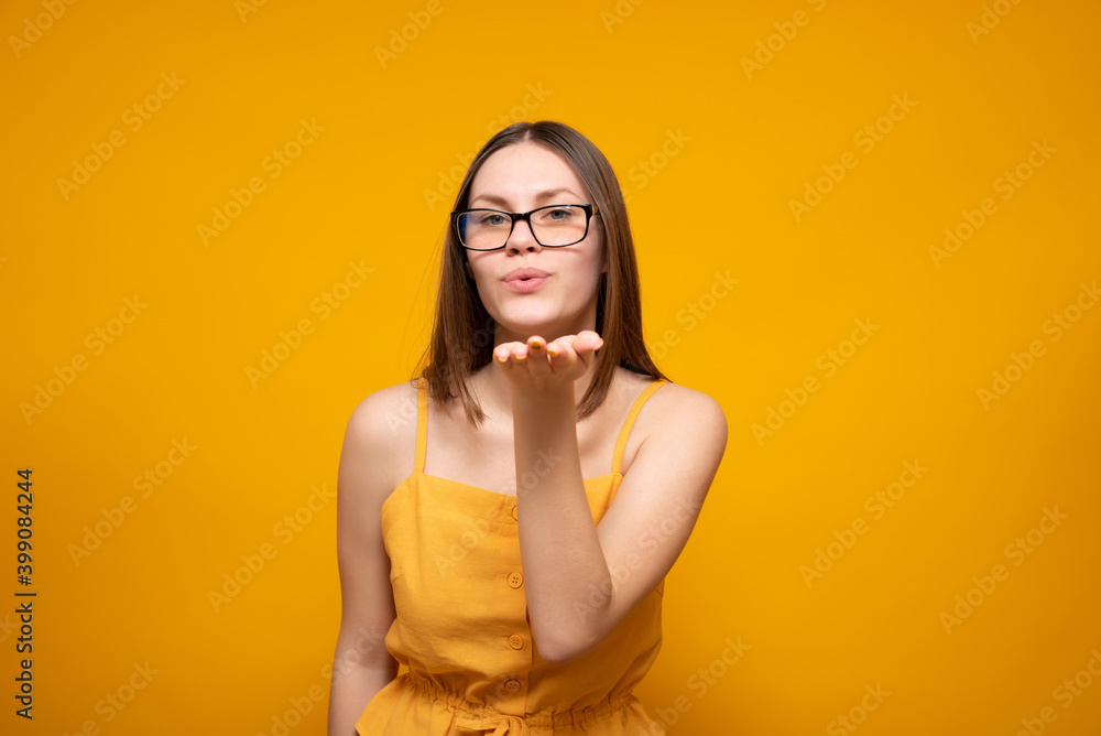 A beautiful young Caucasian woman with glasses blows smb a kiss. isolated on a yellow background. A beautiful young Caucasian woman with glasses blows smb a kiss. isolated on a yellow background.