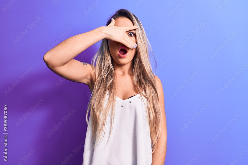 Young beautiful blonde woman wearing white t-shirt standing over isolated purple background peeking in shock covering face and eyes with hand, looking through fingers afraid Young beautiful blonde woman wearing white t-shirt standing over isolated purple background peeking in shock covering face and eyes with hand, looking through fingers afraid