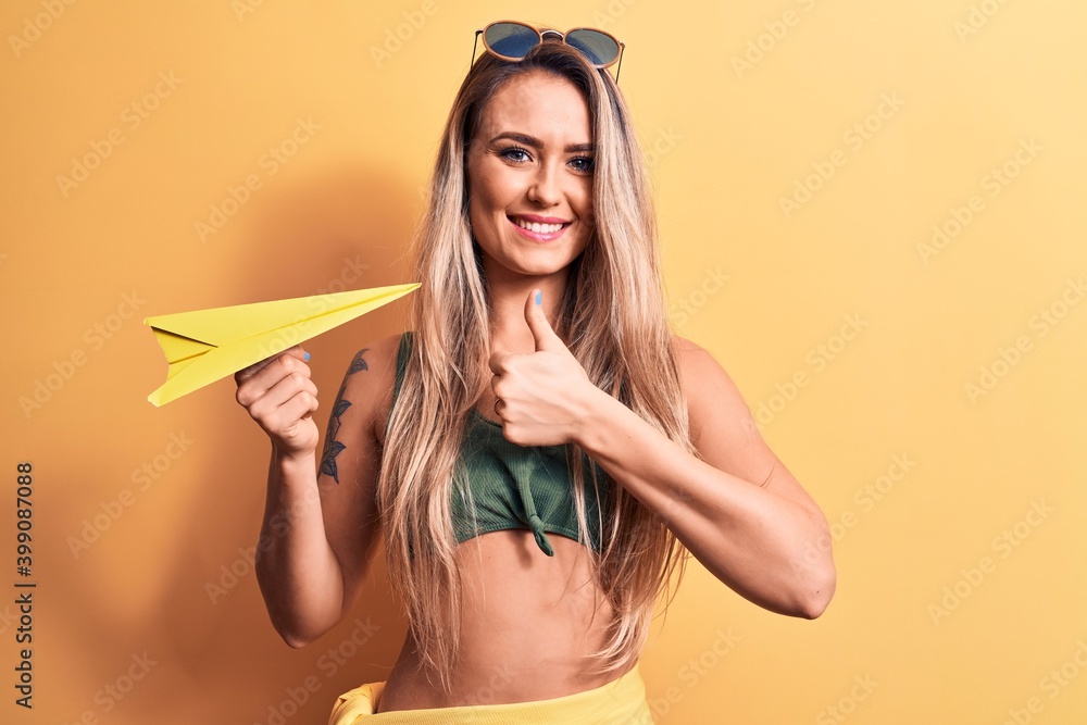 Young beautiful blonde woman wearing bikini and sunglasses holding yellow paper airplane smiling happy and positive, thumb up doing excellent and approval sign