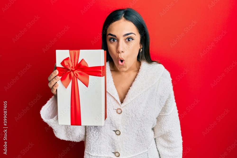 Young brunette woman holding gift scared and amazed with open mouth for surprise, disbelief face Young brunette woman holding gift scared and amazed with open mouth for surprise, disbelief face