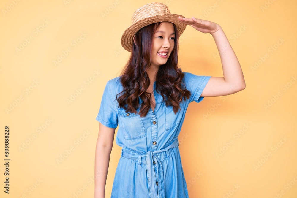 Young beautiful chinese girl wearing summer hat very happy and smiling looking far away with hand over head. searching concept. Young beautiful chinese girl wearing summer hat very happy and smiling looking far away with hand over head. searching concept.