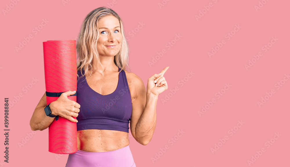 Middle age fit blonde woman holding yoga mat smiling happy pointing with hand and finger to the side Middle age fit blonde woman holding yoga mat smiling happy pointing with hand and finger to the side