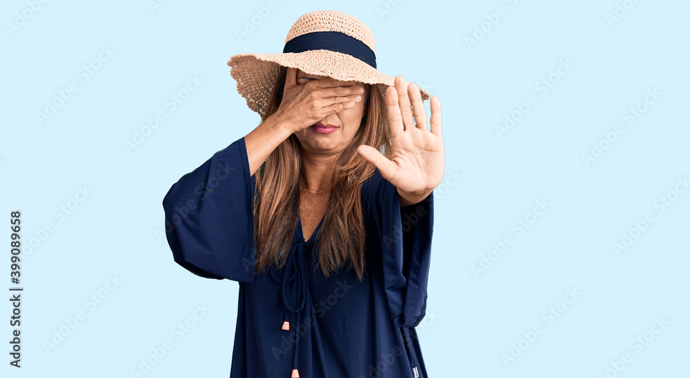 Middle age hispanic woman wearing summer hat covering eyes with hands and doing stop gesture with sad and fear expression. embarrassed and negative concept. Middle age hispanic woman wearing summer hat covering eyes with hands and doing stop gesture with sad and fear expression. embarrassed and negative concept.