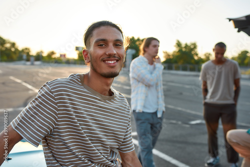 A close up of a casually dressed dark-skinned latina guy with a piercing smiling looking into a camera standing outside on a parking site with his friends on a background