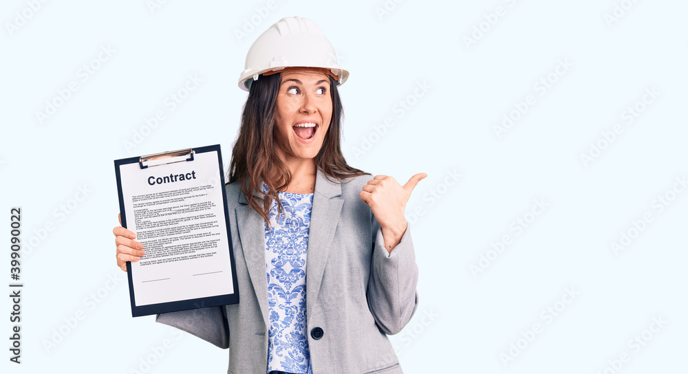 Young beautiful brunette woman wearing architect hardhat holding contract pointing thumb up to the side smiling happy with open mouth Young beautiful brunette woman wearing architect hardhat holding contract pointing thumb up to the side smiling happy with open mouth