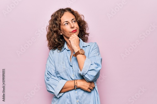 Middle age beautiful woman wearing casual denim shirt standing over pink background with hand on chin thinking about question, pensive expression. Smiling with thoughtful face. Doubt concept.
